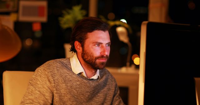 Man is focused and serious while working late on his computer at the office. This image can be used for content related to dedication, professional life, work ethics, business environment, overtime work, and modern workspaces.