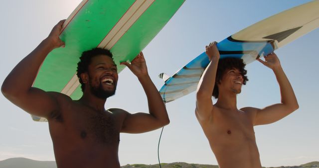 Two young men walking across sandy beach while carrying surfboards on bright sunny day. Both have smiling expressions, enjoying seaside vacation. Perfect for themes like outdoor activities, sports, surfing lifestyle, travel, and summer adventures.
