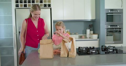 Mother and daughter unpacking groceries at kitchen island with paper bags and oranges