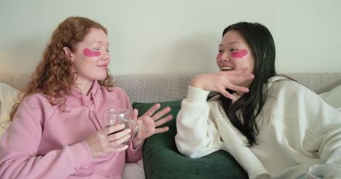 Diverse friends chatting on couch wearing pink under-eye patches enjoying cozy skincare