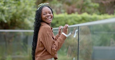 Laughing african american girl leaning on glass balcony railing, braided hair, green bow