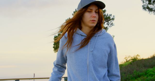 A young Caucasian woman wearing a hoodie and a cap is walking outdoors, with copy space. Her focused expression suggests she might be deep in thought or enjoying a moment of solitude.
