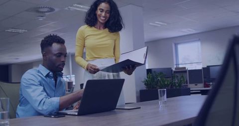 Collaborating over laptop and documents, woman presenting folder to colleague in modern office