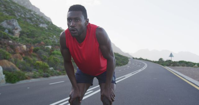 A young athletic man is taking a short rest break during a run on a scenic mountain road. He is wearing a red sports shirt and black workout shorts. This picture captures the effort and determination involved in athletic activities. Ideal for use in fitness, health, and wellness promotions, as well as social media campaigns focused on outdoor activities or sports motivation.