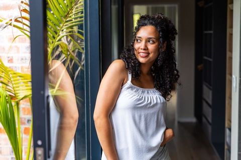 African american woman leaning by glass door, smiling and relaxing in sunlit minimalist home