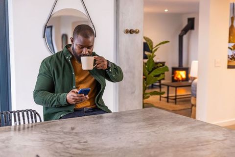 African american man sipping coffee and checking phone at modern kitchen island by fireplace