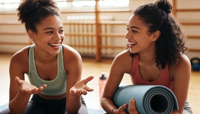 Laughing teens chatting after workout, holding yoga mat in bright fitness studio