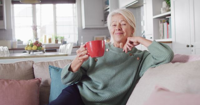 Portrait of happy senior caucasian woman sitting on sofa in living room, drinking coffee. Spending quality time at home alone.