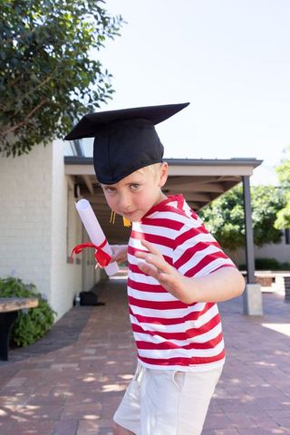 Playful 6-year-old boy wearing graduation cap holding diploma celebrating milestone