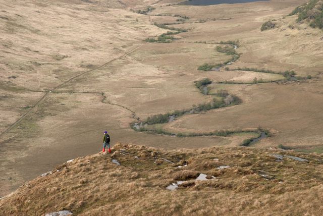 Single hiker standing on top of a hill, overlooking a vast and remote valley with a winding river below. Ideal for use in travel articles, outdoors and adventure blogs, and promotional material for hiking trails or countryside retreats. Captures feelings of serenity, exploration, and the beauty of nature.
