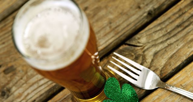 A glass of beer is placed next to a fork and a green shamrock, symbolizing a festive meal for St. Patrick's Day. The wooden table background adds a rustic charm to the celebration of this Irish holiday.