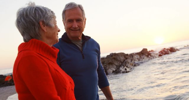 Senior couple walking along ocean shore during sunset, smiling and enjoying each other's company. Perfect for illustrating concepts of aging gracefully, retirement lifestyle, and romantic beach scenery. Ideal for use in travel brochures, retirement planning advertisements, healthy living campaigns, or any media focusing on elderly wellness and happiness.