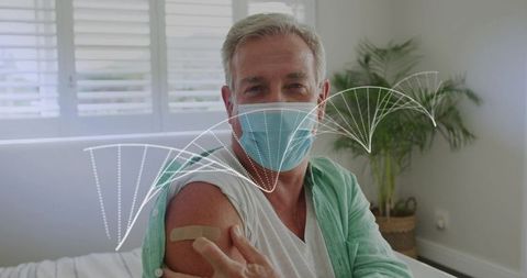Senior man showing vaccination bandage on upper arm wearing face mask in sunlit bedroom