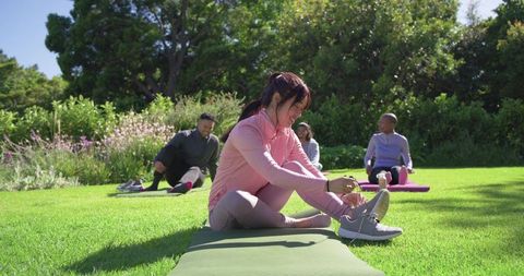 Young asian woman tying running shoe on yoga mat in park during group outdoor workout