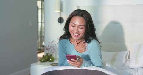 Mid adult asian woman smiling while texting on smartphone in cozy bedroom morning light