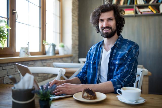 Man with beard using laptop in coffee shop, smiling at camera. Natural light from window, casual setting with coffee and pastry on table. Ideal for themes of remote work, freelancing, modern lifestyle, technology, and casual work environments.