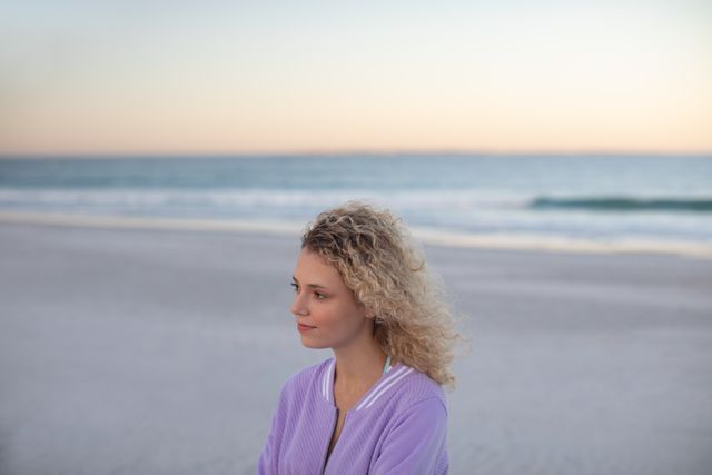 This image captures a thoughtful woman with curly hair standing on a beach at sunset. The serene ocean and horizon in the background create a peaceful atmosphere. Ideal for use in mental health, wellness, and travel content, as well as advertisements promoting relaxation and introspection.
