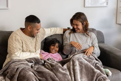 Diverse family laughing and cuddling under plush blanket on gray sofa, cozy home moment