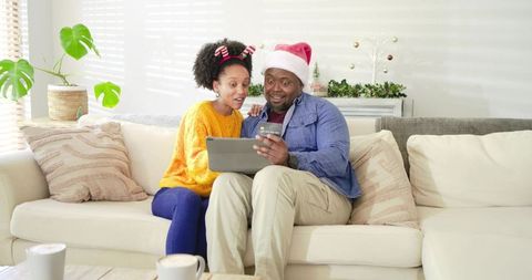 Couple shopping online for holiday gifts on tablet while wearing santa hats and cozy sweaters