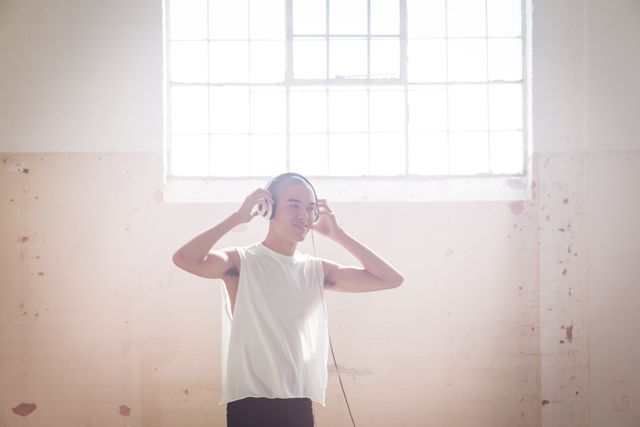 Front view of a hip young biracial man in an empty warehouse, wearing headphones and holding them, smiling, backlit by sunlight by the window.