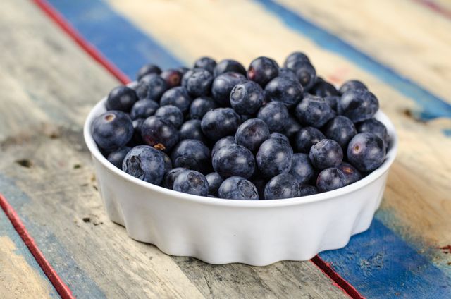 Fresh blueberries are displayed in a white bowl resting on a rustic wooden table painted with blue and red stripes. The vibrant color of the ripe blueberries contrasts with the distressed wood, emphasizing their natural and appetizing look. This image is perfect for promoting healthy eating, organic produce, and nutrition-related content. It can be used for cookbooks, food blogs, restaurant menus, or advertisements for fresh fruit.