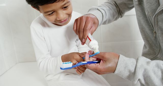 A parent helps a young child apply toothpaste to a toothbrush in a bathroom. This image can be used to promote family bonding, dental health awareness, or products related to oral hygiene. Suitable for use in parenting magazines, dental care advertisements, and healthcare brochures.