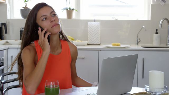 Young woman wearing orange sleeveless top using mobile phone and working on a laptop while sitting at dining table in a modern kitchen. Green smoothie in glass next to her. Could be used for themes related to remote work, home office, multitasking, modern lifestyle, or technology in everyday life.