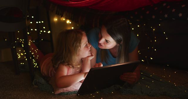 A mother and daughter are lying under a blanket fort, reading from a tablet surrounded by warm fairy lights. This image is perfect for showcasing family bonding, cozy home moments, and parenting. It can be used in advertisements for family products, digital books, and parenting blogs.