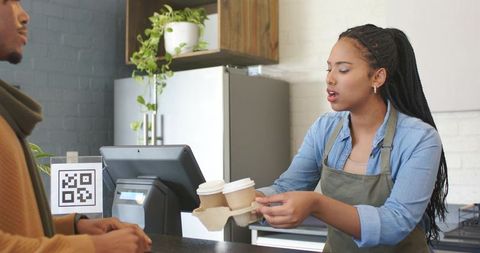 Young female barista handing coffee carrier to african american man at modern cafe counter