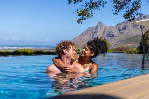 Romantic couple embracing, smiling in infinity pool with ocean horizon and mountain view