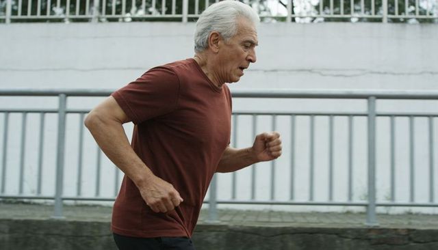 Active senior man jogging along urban sidewalk in maroon shirt fitness and determination