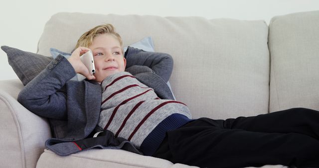 Young boy in casual wear is lying on the couch, talking on a smartphone and relaxing. This image can be used for advertisements featuring children-friendly technology, modern communication for kids, or family lifestyle.