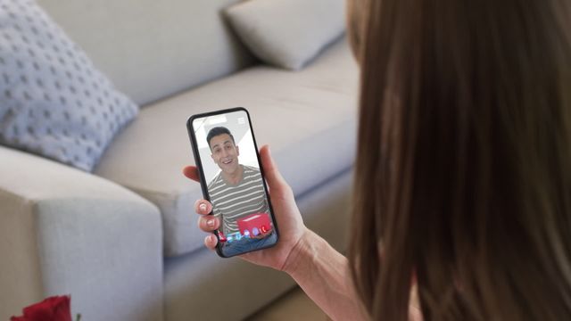 Caucasian woman holding a red rose while video calling her boyfriend on a smartphone shows a mixed-race man smiling with a gift. Perfect visual for themes of long-distance relationships, virtual dating, and celebrating Valentine's Day with technology.