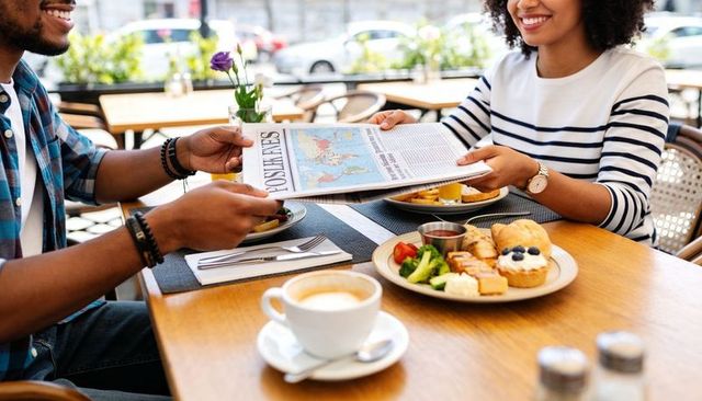Couple passing newspaper over brunch at outdoor cafe terrace, sharing coffee and pastries