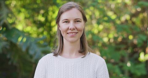 Serene woman in white cable-knit sweater smiling in sunlit garden with soft bokeh, soft light
