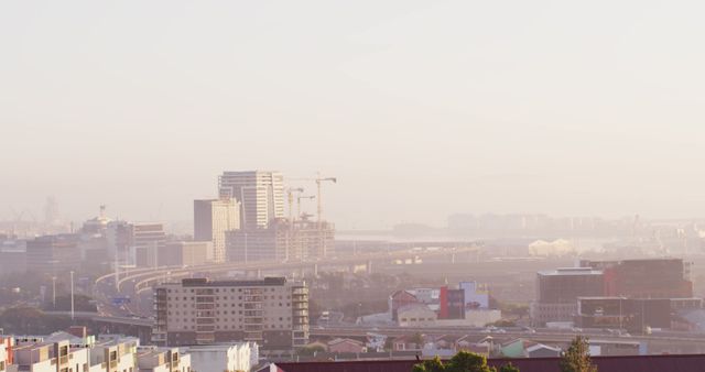 Urban landscape showing city skyline covered in fog during sunrise. Buildings and construction cranes visible in the distance with parts of the harbor also in view. This image is perfect for illustrating morning city life, urban development, and atmospheric conditions. Can be used in travel brochures, weather reports, and real estate promotions.