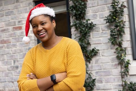 African american woman wearing santa hat, mustard sweater, smiling and crossing arms outdoors