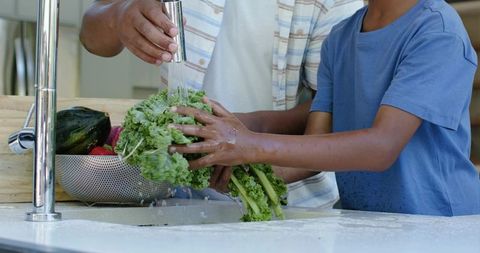 African american father and son washing kale together in kitchen teaching healthy cooking