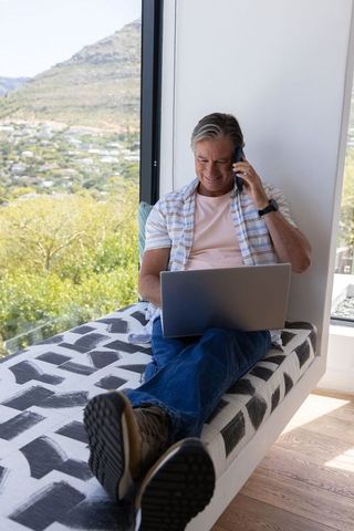 Middle-aged man working remotely on laptop and phone in window seat with hillside view