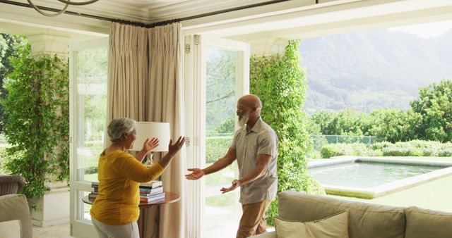 Senior couple in modern living room extending hands for embrace. Open doors reveal greenery and mountains with a pool outside. Suitable for depicting retirement lifestyle, love, happiness in mature relationships, and advertisements for home living spaces and family life.