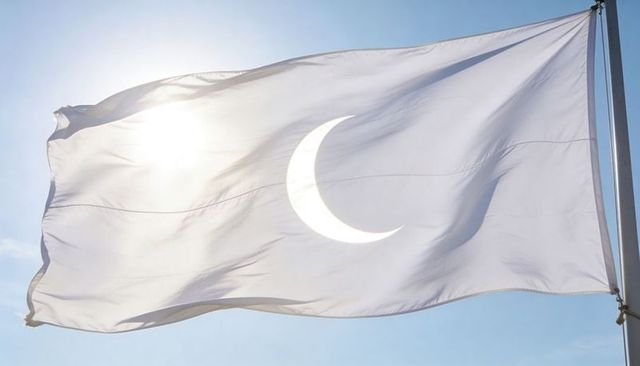 Backlit white flag with crescent emblem waving against clear blue sky and sun flare