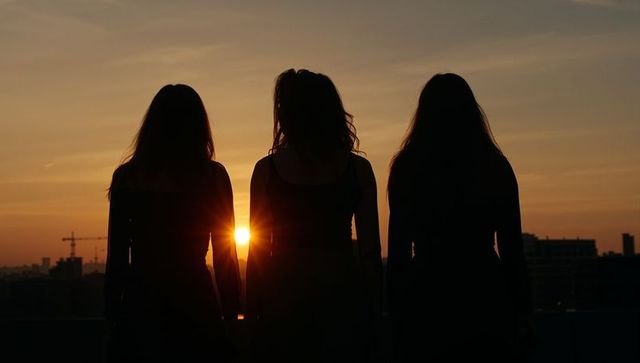 Three female silhouettes watching urban sunset on rooftop with city skyline and crane