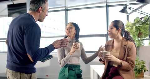 Diverse Coworkers Chatting Over Coffee in Bright Modern Office with Urban Views, Casual Meeting