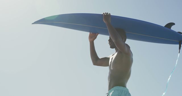 Happy african american man carrying surfboard on head walking on sunny beach by the sea. Summer, hobbies, surfing and vacations.