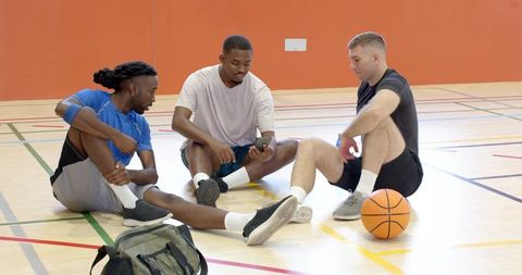 Three male teammates sitting on indoor basketball court sharing phone after pickup game