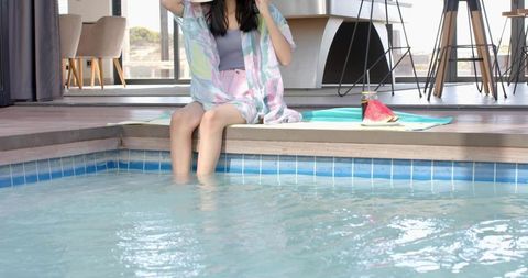 Relaxing by pool in pastel cover-up dipping legs with sun hat and watermelon snack
