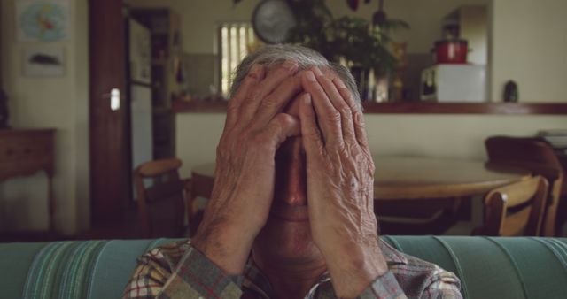 Elderly man sitting alone with hands covering his face, possibly feeling frustrated or worried. Man seated on sofa in living room or dining area. Useful for themes related to senior mental health, solitude, retirement, aging, anxiety management, and offering supportive services for the elderly.