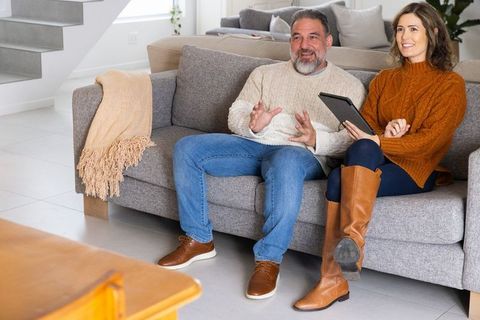 Middle-aged couple sitting on sofa in minimalist living room using tablet while smiling