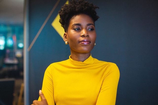 Confident African American businesswoman wearing yellow top, looking away in modern office. Ideal for use in corporate websites, business presentations, career development materials, and diversity and inclusion campaigns.