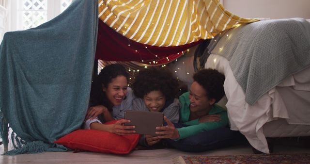 Friends enjoying time together under a cozy blanket fort, laughing and using a tablet. Ideal for depicting friendship, indoor activities, and leisure moments at home. Suitable for promoting tech gadgets, family activities, and lifestyle blogs focusing on diverse cultural experiences.